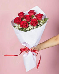 6 red roses and baby's breath wrapped in white tissue paper with a thin red ribbon. The bouquet is handheld against a pink background.