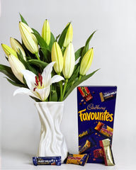 Bouquet of white and yellow lilies in a white vase next to a box of Cadbury Favourites on a light gray background