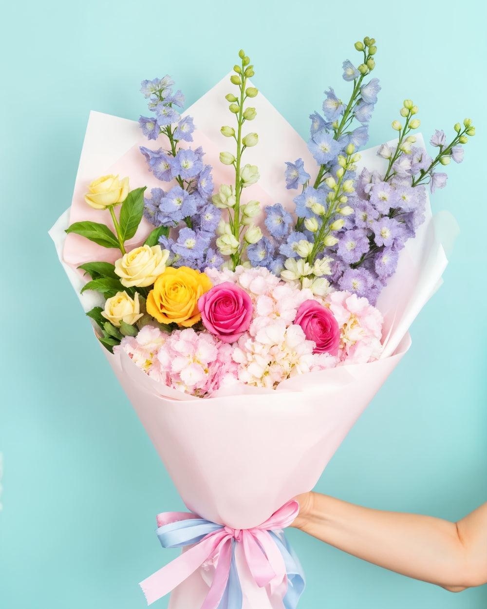 Pastel Bouquet on a blue background. The bouquet consists of 2 hydrangeas, 5 snapdragons and 6 roses. The bouquet is handheld and includes a pink and blue ribbon.