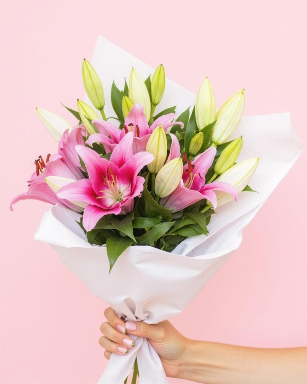 18 pink lilies wrapped in white tissue paper and handheld against aa soft pink background.