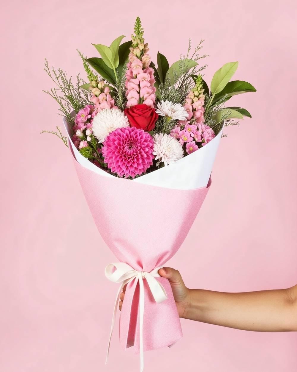 Small pink posy of flowers featuring red, pink and white flowers. The bouquet is handheld on a pink background and wrapped in pink tissue paper with a white ribbon.