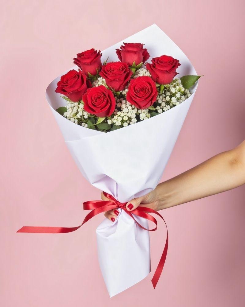 6 red roses and baby's breath wrapped in white tissue paper with a thin red ribbon. The bouquet is handheld against a pink background.