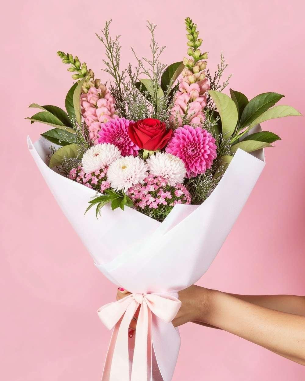 A romantic flower bouquet consisting of assorted pink, red and white flowers with greenery. The flowers are wrapped in white tissue paper with a pink ribbon.